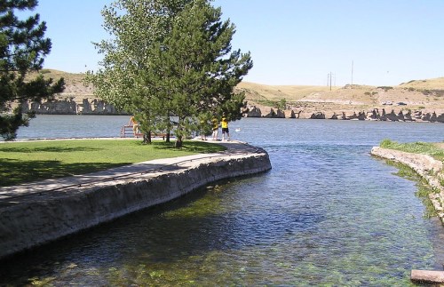 A serene river scene with a grassy bank, trees, and people enjoying the outdoors on a sunny day.
