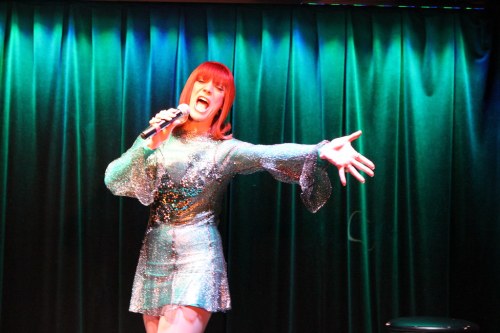 A performer in a sparkly silver dress sings passionately on stage under green lighting.