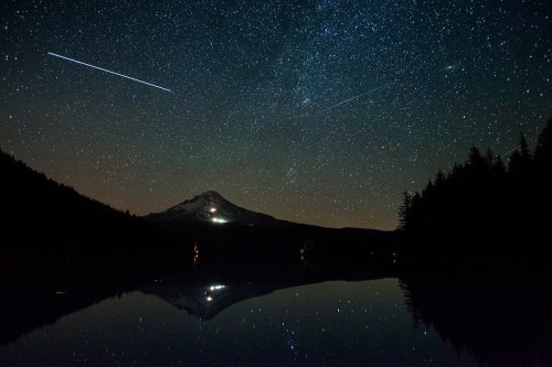 A starry night sky over a mountain reflecting in a calm lake, with trees framing the scene and shooting stars above.