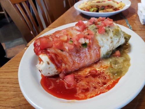A large burrito topped with red and green sauces, diced tomatoes, served on a white plate with a wooden table background.