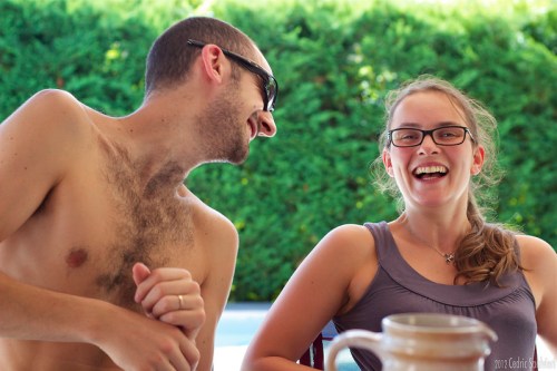 A man and woman laugh together outdoors, enjoying a sunny day by a pool, with greenery in the background.