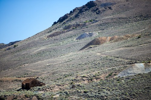 A weathered wooden cabin sits on a barren hillside, surrounded by sparse vegetation and mining debris.
