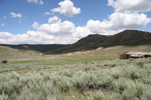 A vast landscape featuring rolling hills, green sagebrush, and scattered clouds in a blue sky. Abandoned structures are visible.