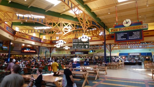 A bustling food court with colorful decor, wooden tables, and a variety of food stalls filled with people.