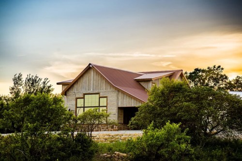 A modern wooden house with a metal roof, surrounded by greenery, under a colorful sunset sky.