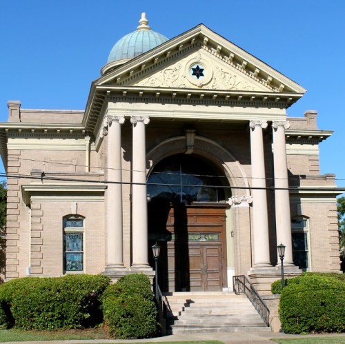 Historic building with a dome, columns, and a large entrance, surrounded by greenery and a clear blue sky.