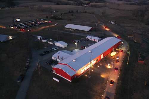 Aerial view of a red barn-like building surrounded by cars, with fields and a parking lot illuminated at dusk.