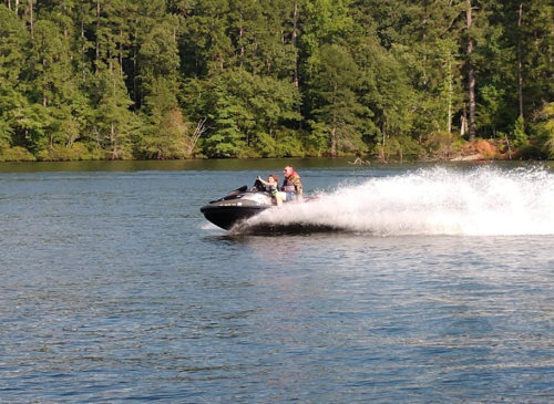 A person rides a jet ski across a lake, creating a spray of water against a backdrop of lush green trees.