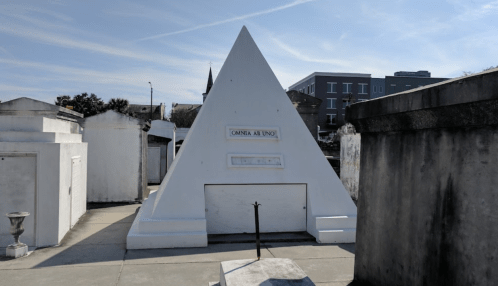 A white pyramid-shaped tomb in a cemetery, surrounded by other mausoleums and buildings under a clear blue sky.