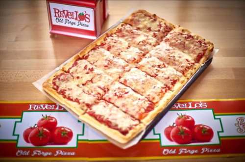 A rectangular pizza with melted cheese and tomato sauce, placed on a wooden surface next to a branded box and banner.