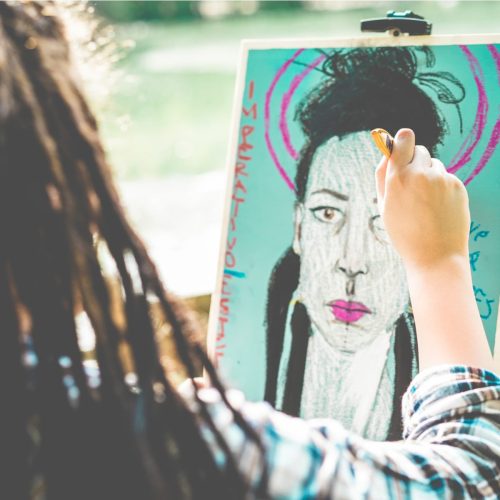 A person drawing a colorful portrait of a woman with a serious expression, surrounded by greenery.