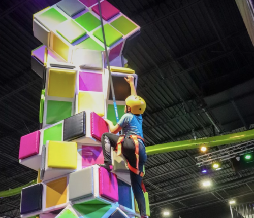 A child in a helmet climbs a colorful indoor climbing wall, reaching for the top with a safety harness attached.