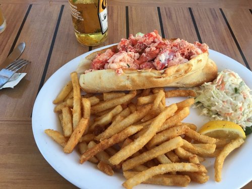 A plate with a lobster roll, fries, coleslaw, and a lemon wedge, alongside a bottle of beer on a wooden table.