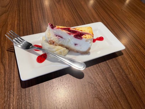 A slice of cheesecake topped with raspberry sauce on a white plate, accompanied by a fork, on a wooden table.