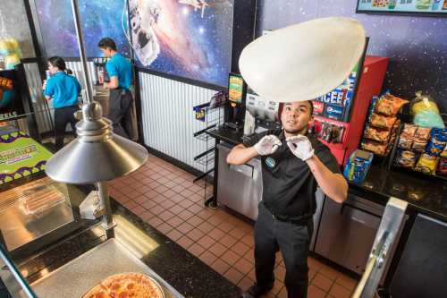 A staff member tosses pizza dough in a colorful pizzeria with a space-themed mural in the background.