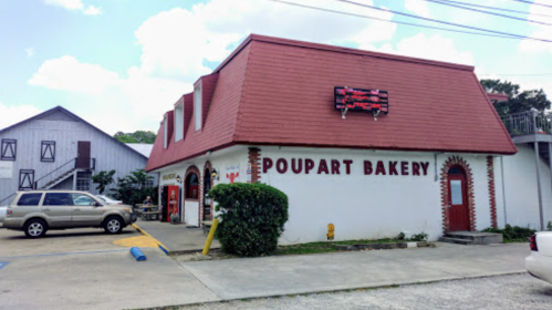 A bakery with a red roof and sign, surrounded by greenery and parked cars, under a blue sky with clouds.
