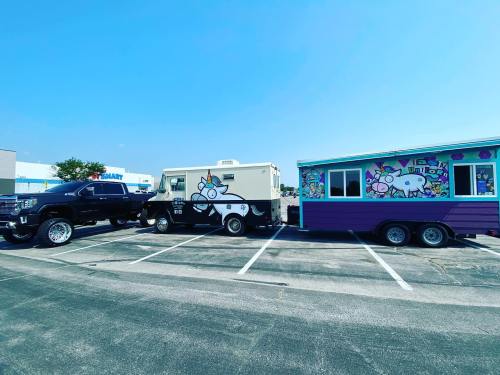 A food truck and a colorful trailer parked in a lot, with a black truck beside them under a clear blue sky.