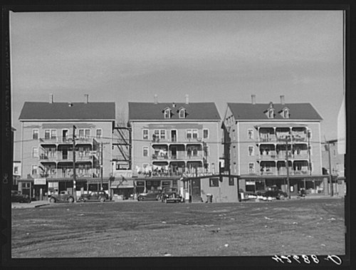 Three multi-story apartment buildings with balconies, set against a clear sky, with parked cars in the foreground.