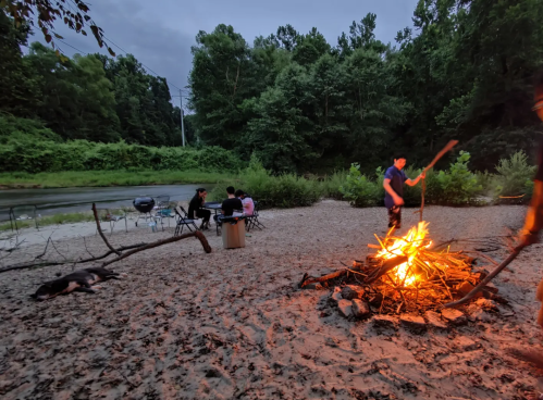 A campfire burns on a sandy beach by a river, with people sitting nearby and trees in the background.
