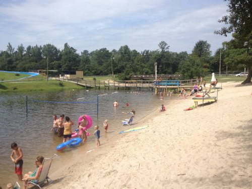 A sandy beach area with children and adults swimming, playing, and relaxing by a calm lake under a partly cloudy sky.