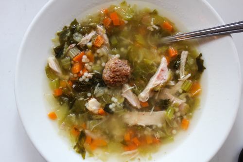 A bowl of soup with greens, carrots, chicken, and a meatball, served with a spoon.