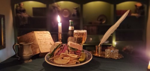 A plate of sandwiches and chips beside a candle, beer, and vintage decor, creating a cozy dining atmosphere.