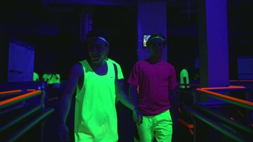 Two individuals in neon athletic wear walk through a dimly lit space with glowing accents.