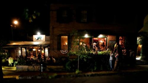 A cozy restaurant at night, with warm lights, outdoor seating, and people socializing outside.