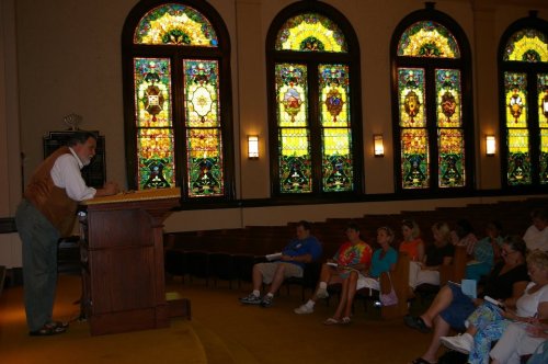 A speaker at a podium addresses an audience in a room with colorful stained glass windows.