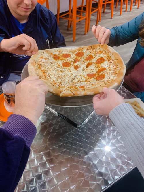 Four hands reaching for a large pepperoni pizza on a metal tray at a casual dining setting.