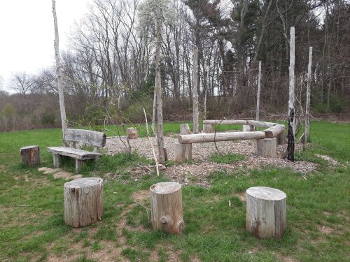 A rustic gathering area with log benches and a circular arrangement of wooden posts, surrounded by grass and trees.