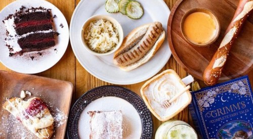 A table spread with various dishes: cake, a sandwich, pickles, sauerkraut, pretzel, and a book.