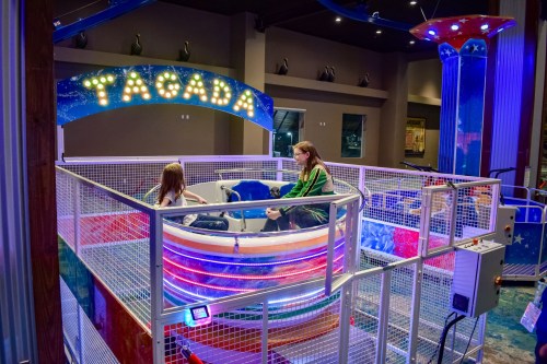 Two girls sit in a colorful amusement ride surrounded by bright lights and a mesh enclosure.