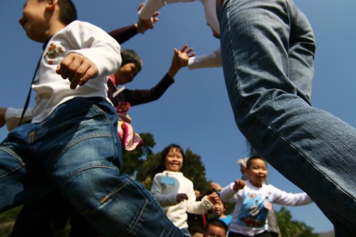 A group of children and adults play together outdoors, reaching out and smiling under a clear blue sky.