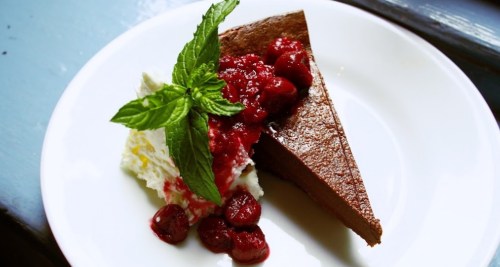 A slice of chocolate cake topped with raspberry sauce, whipped cream, and a sprig of mint on a white plate.