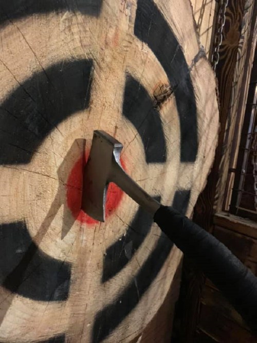 An axe embedded in a wooden target with a red bullseye and black concentric circles.