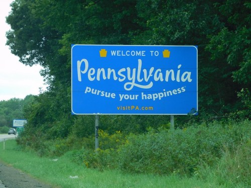 Blue welcome sign for Pennsylvania with the slogan "pursue your happiness" and a website link at the bottom.