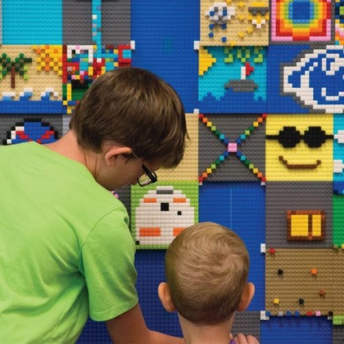 Two children create a colorful mural with LEGO bricks on a blue wall, focused on their artwork.