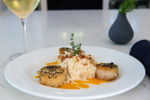 A plate of seared scallops on creamy risotto, garnished with herbs and a drizzle of sauce, with a glass of white wine nearby.