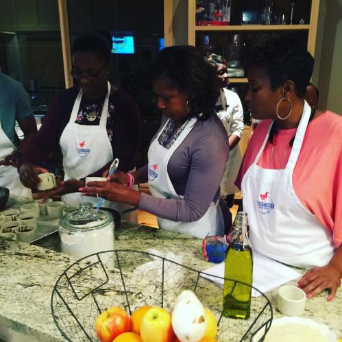 A group of women in aprons preparing food in a kitchen, focused on measuring ingredients. Apples and oil are on the counter.