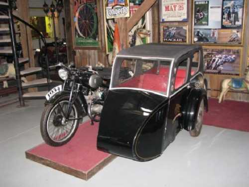 A vintage black car with a red interior next to a classic motorcycle, displayed in a rustic museum setting.