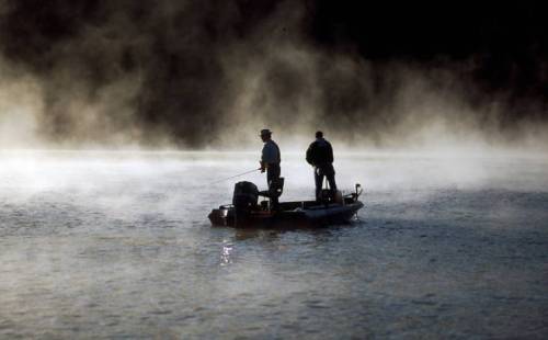 Two fishermen on a boat in a misty lake at dawn, casting lines into the calm water.