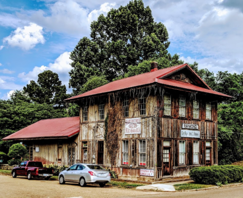A rustic, two-story wooden building with a red roof, surrounded by trees and parked cars on a sunny day.