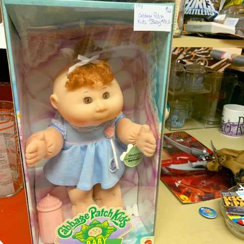 A Cabbage Patch Kids Baby doll in a blue dress, displayed in a box with a pink bottle beside it.