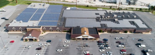 Aerial view of a large building with solar panels on the roof and a parking lot filled with cars.