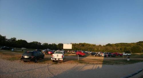 A wide view of a parking lot filled with cars, with a large outdoor screen in the background and trees lining the area.