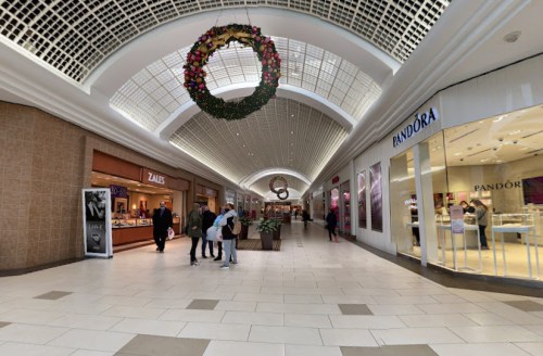 A spacious mall corridor with shops on either side, decorated with a holiday wreath overhead. Shoppers are present.