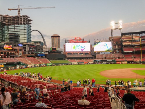 View of a baseball stadium with a large screen, crowds gathering, and city skyline in the background at sunset.