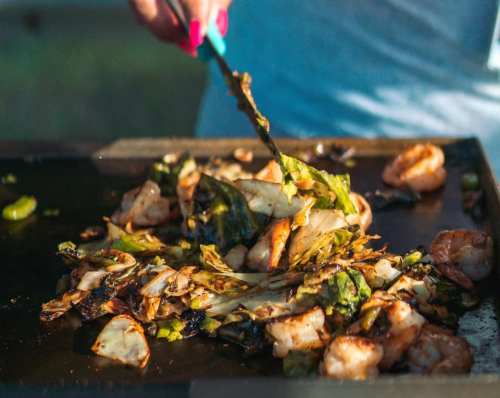 A person using tongs to stir-fry shrimp and vegetables on a grill, with vibrant colors and a sunny outdoor setting.