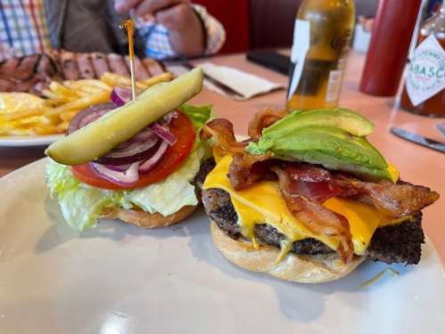 A close-up of a cheeseburger with bacon and avocado, served with a side of fries and a drink in the background.